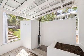 Een overdekt buitengebied met een inbouwdouche, een raam en uitzicht op een huis en bomen.
