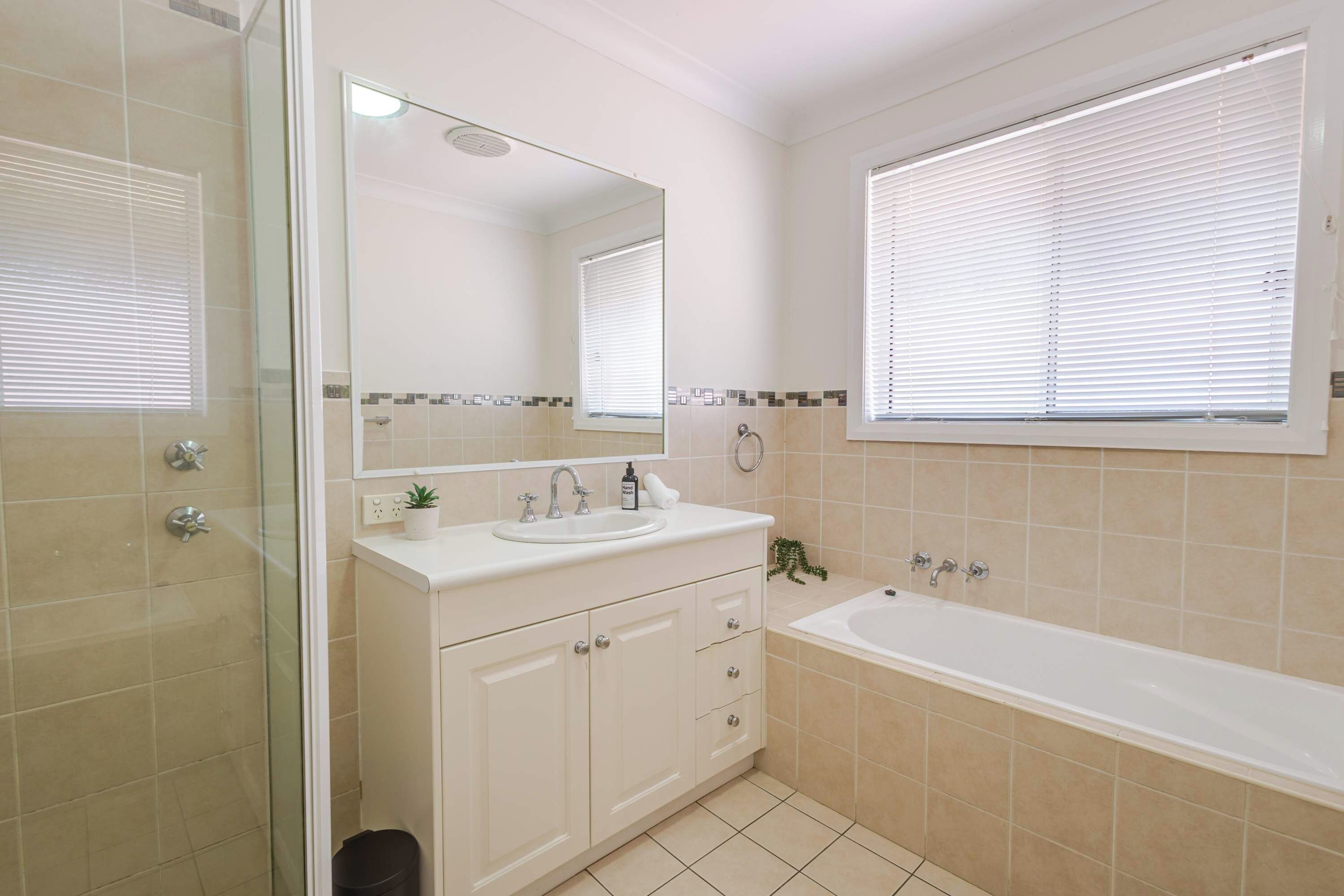 A bathroom with a glass shower enclosure, a sink with a mirror above, a bathtub, and a window with blinds.