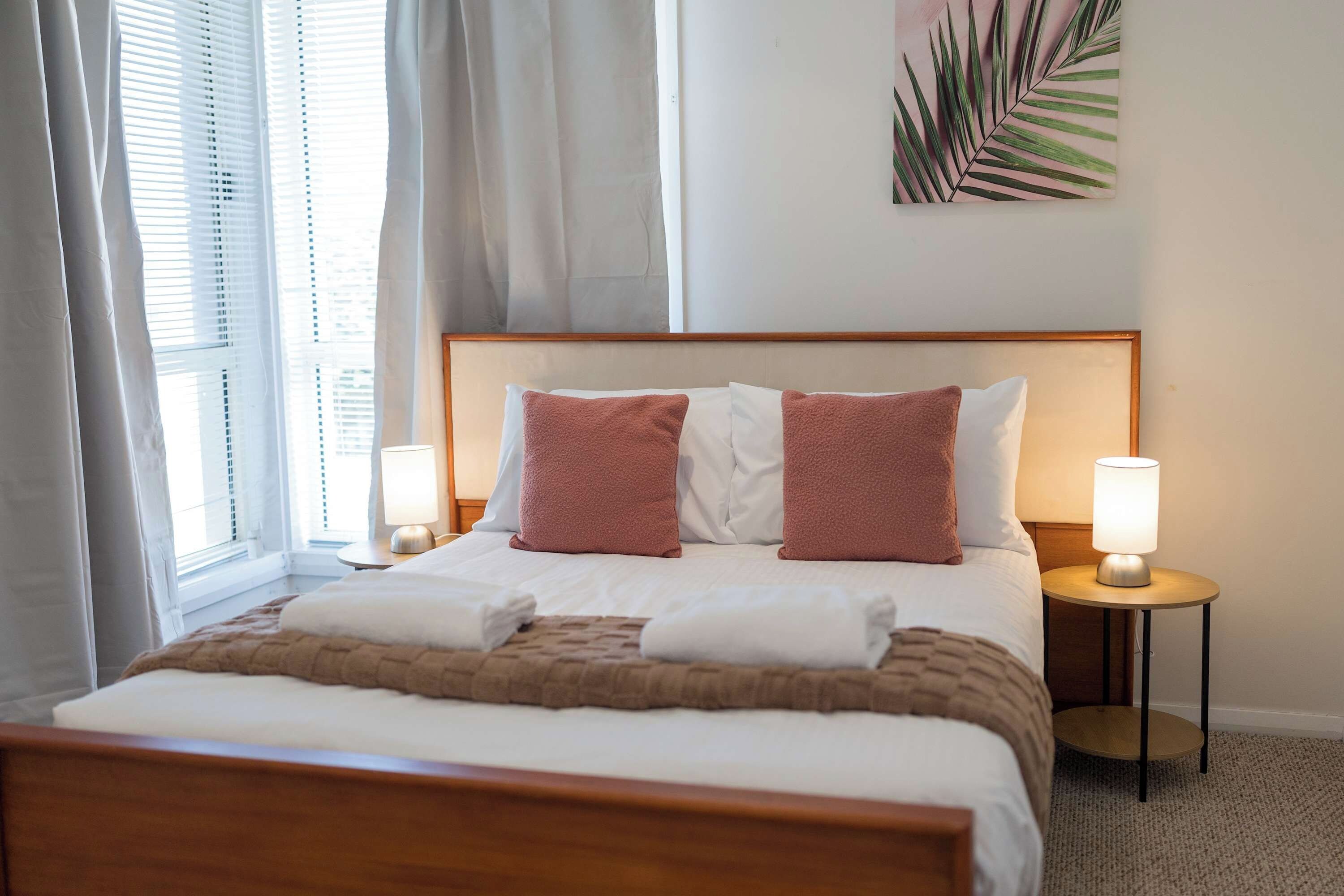 A hotel room with a bed, two bedside lamps, a window with curtains, and a wall-mounted plant.