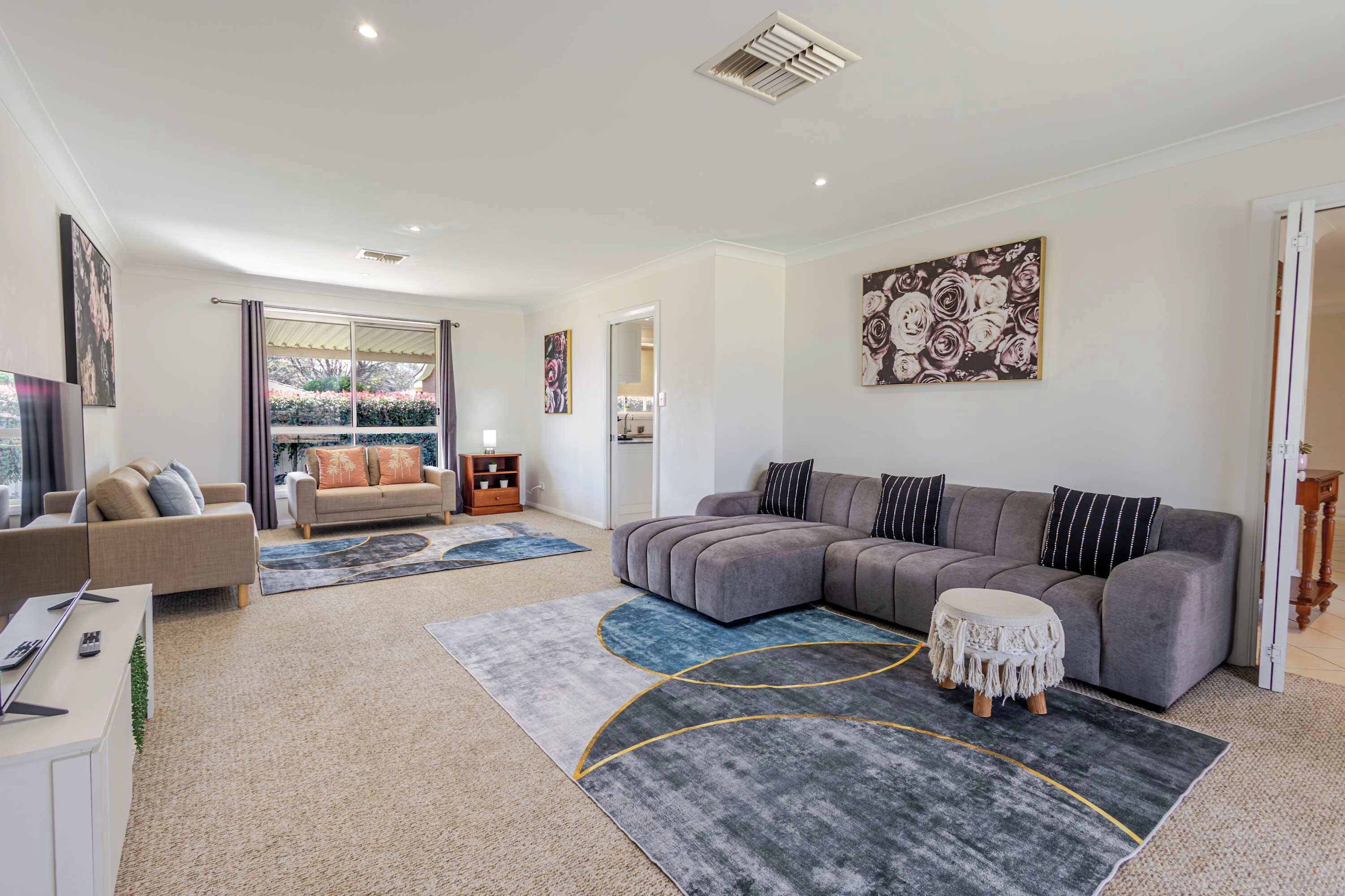 A modern living room with a sectional sofa, a coffee table, and a TV. There are two side tables and a small round table. The room has a large window with curtains, a ceiling light, and a painting on the wall.