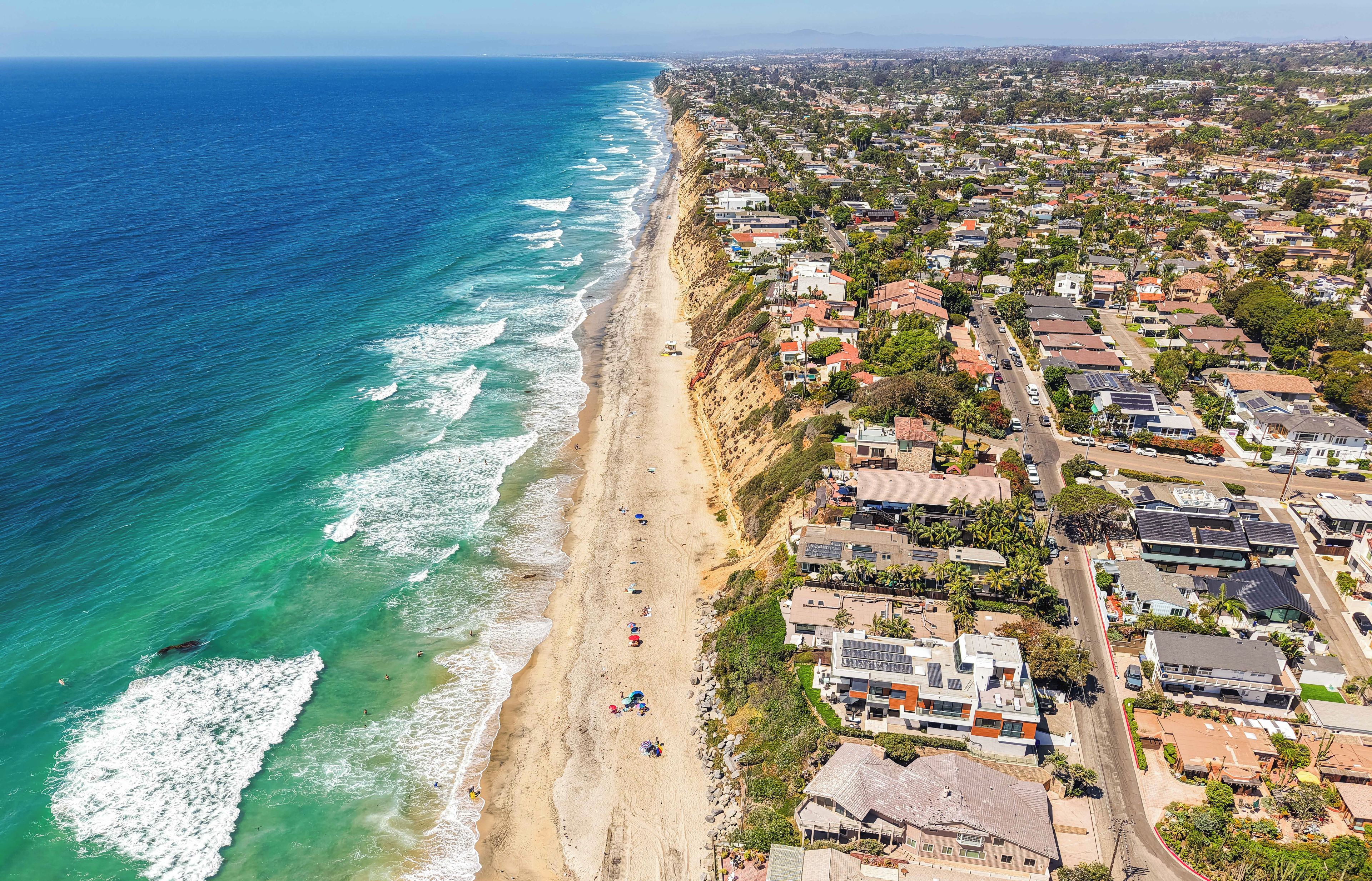 An aerial view of a coastal residential area with houses, a beach, and a clear view of the ocean.