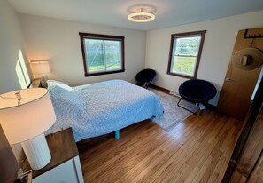 A bedroom with a bed, two windows, a chair, a lamp, and a door with a hat hanging on it. - Get the break you deserve, at the River Respite. (Memphis)