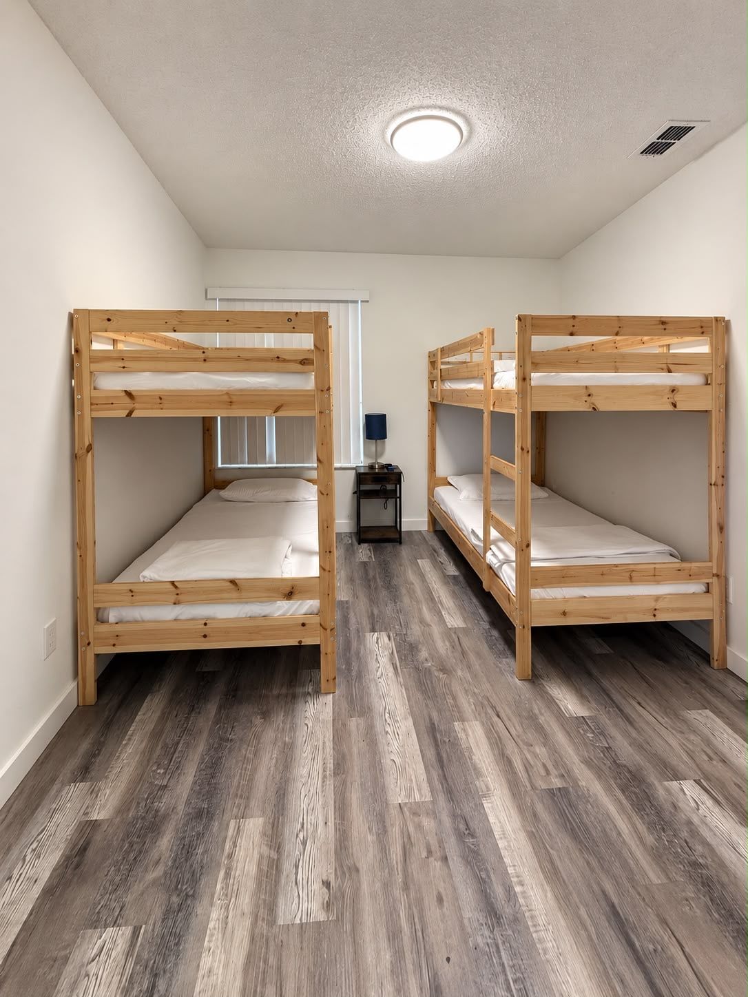 Une chambre avec deux lits superposés, une petite table avec une lampe et une fenêtre avec des stores.