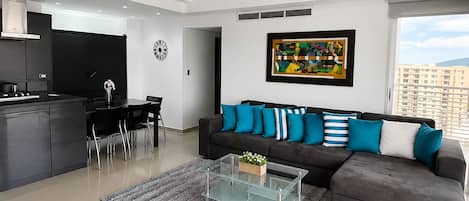 A modern living room with a grey sofa, a glass coffee table, and a dining area with chairs.