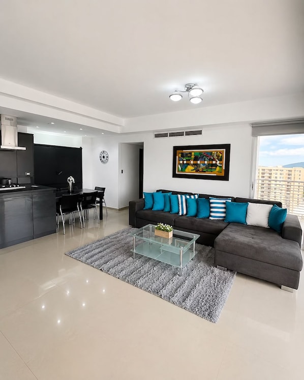 A modern living room with a grey sofa, a glass coffee table, and a dining area with chairs.