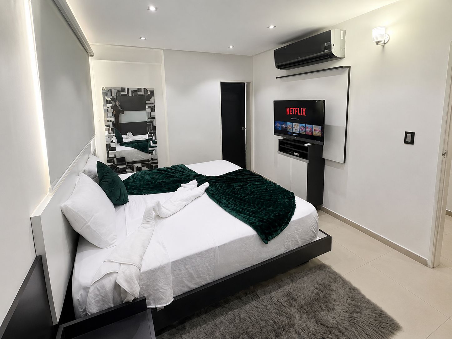 A modern bedroom with a bed, a flat-screen TV mounted on the wall, and a built-in air conditioning unit.