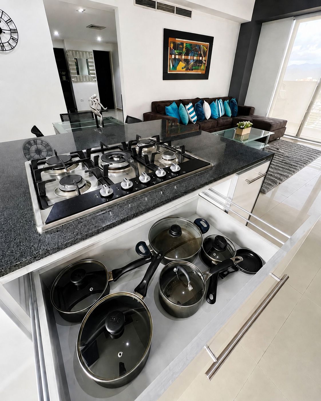 Une cuisine moderne dotée d’une cuisinière à gaz intégrée, d’un îlot central avec une plaque de cuisson en verre et de divers ustensiles de cuisine rangés dans les meubles situés en dessous.
