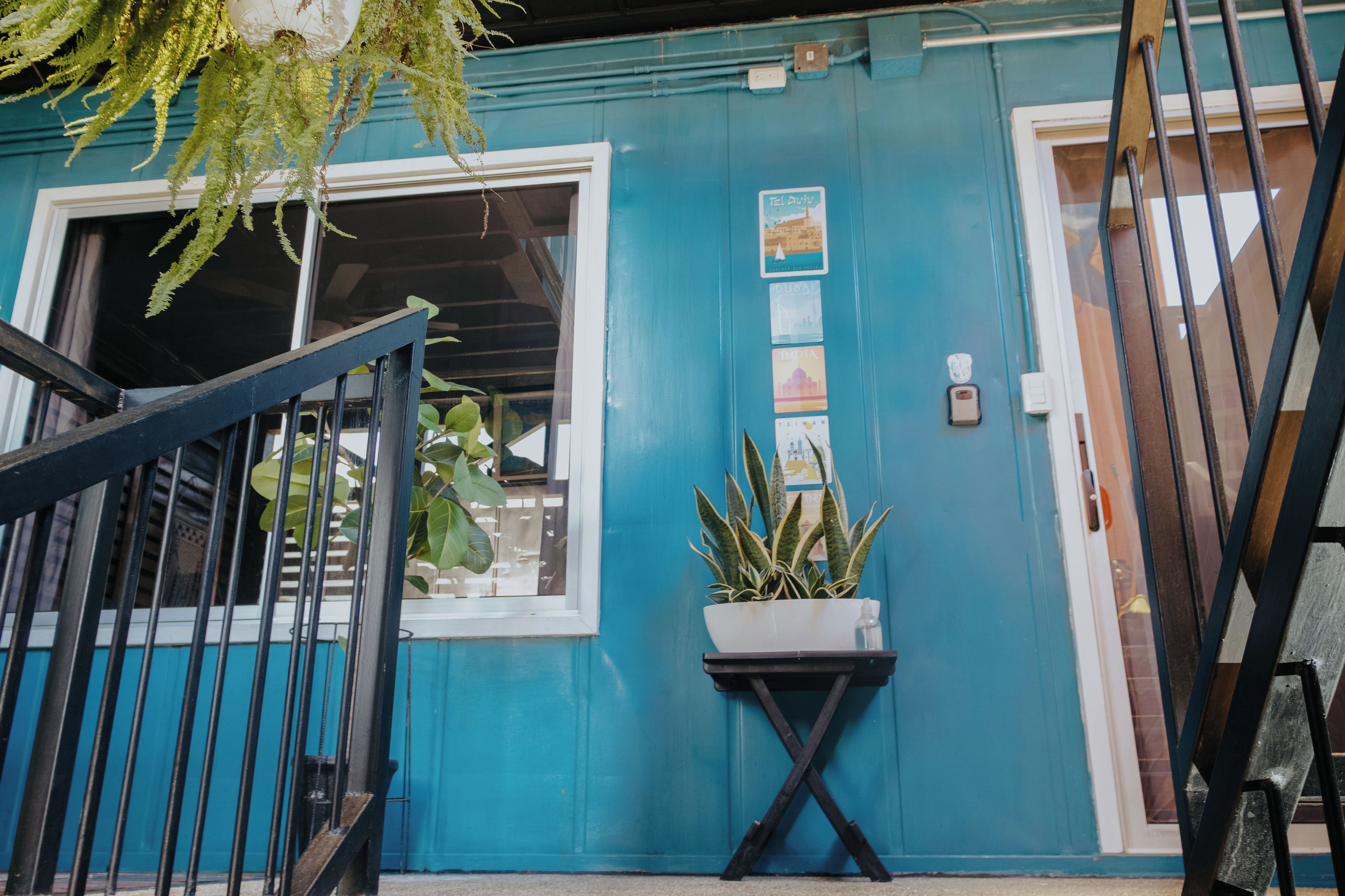 A blue building with a white-framed window, a potted plant on a small table, and a metal gate.