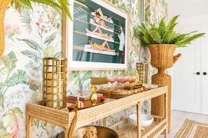 Une pièce décorée d'un papier peint aux motifs tropicaux, une console en osier sur laquelle sont disposées des boissons, et une grande plante en pot.
