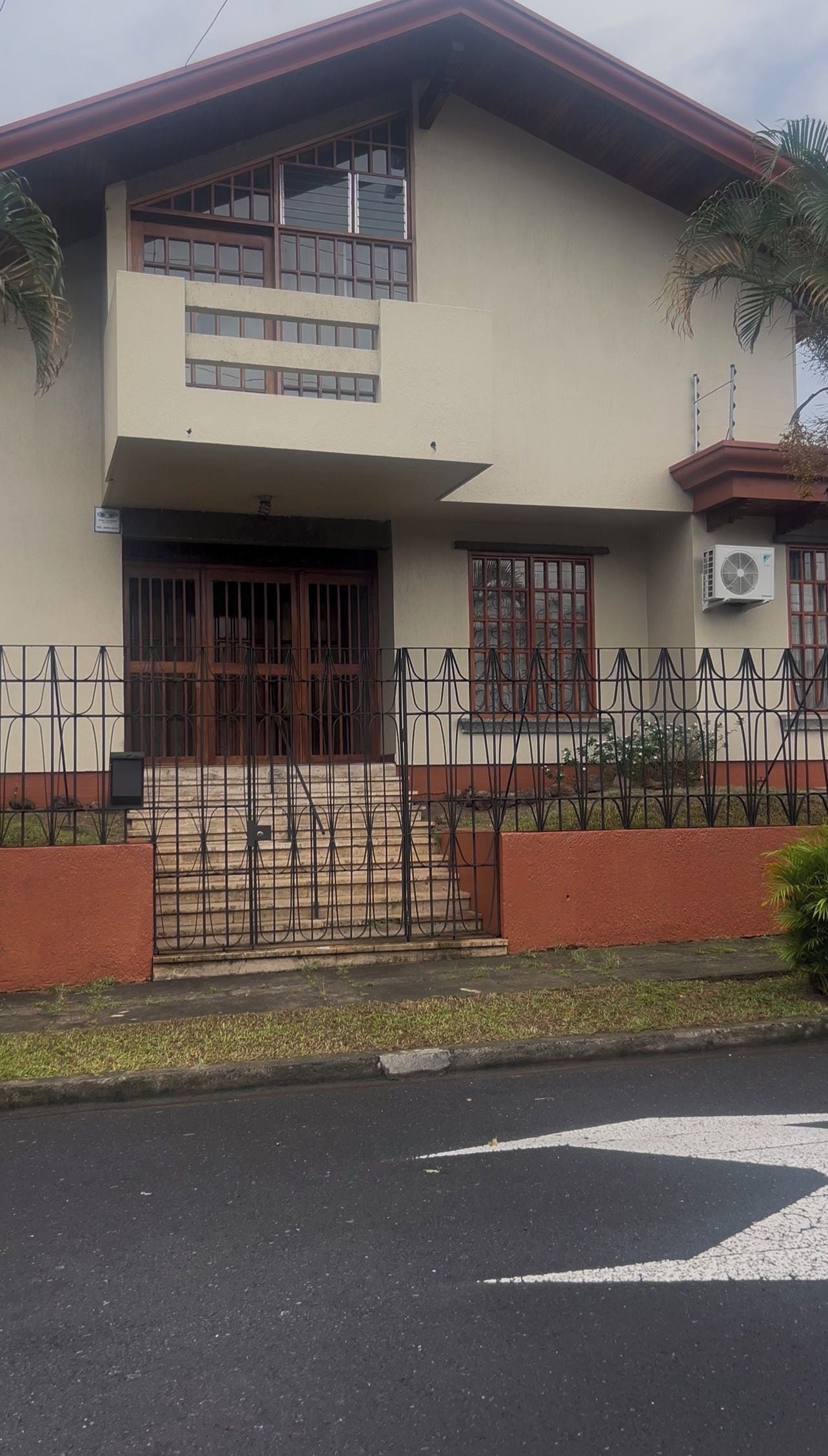Un edificio de dos plantas con entrada protegida por una reja, un pequeño porche y una unidad de aire acondicionado visible.