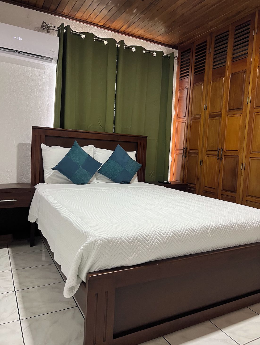 Una cama con cabecera de madera, dos almohadas, una colcha blanca, una mesita de noche y una cortina verde.