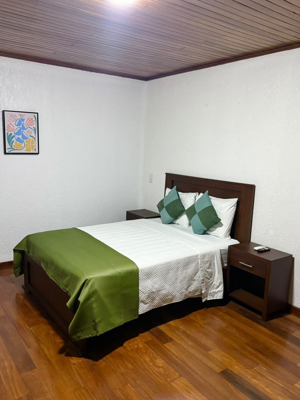 Un lit bien fait, avec une couvre-lit verte et du linge de lit blanc, une tête de lit en bois et une table de chevet sur laquelle se trouve une télécommande.