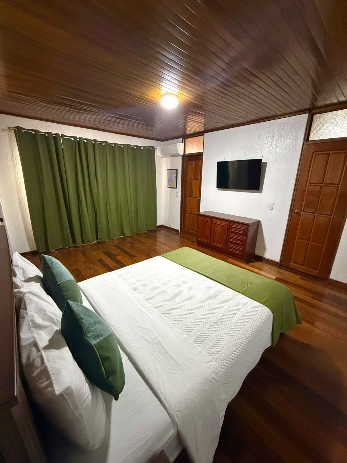 Un dormitorio con cama, cortinas verdes, suelo de madera, televisor en la pared y un armario de madera.