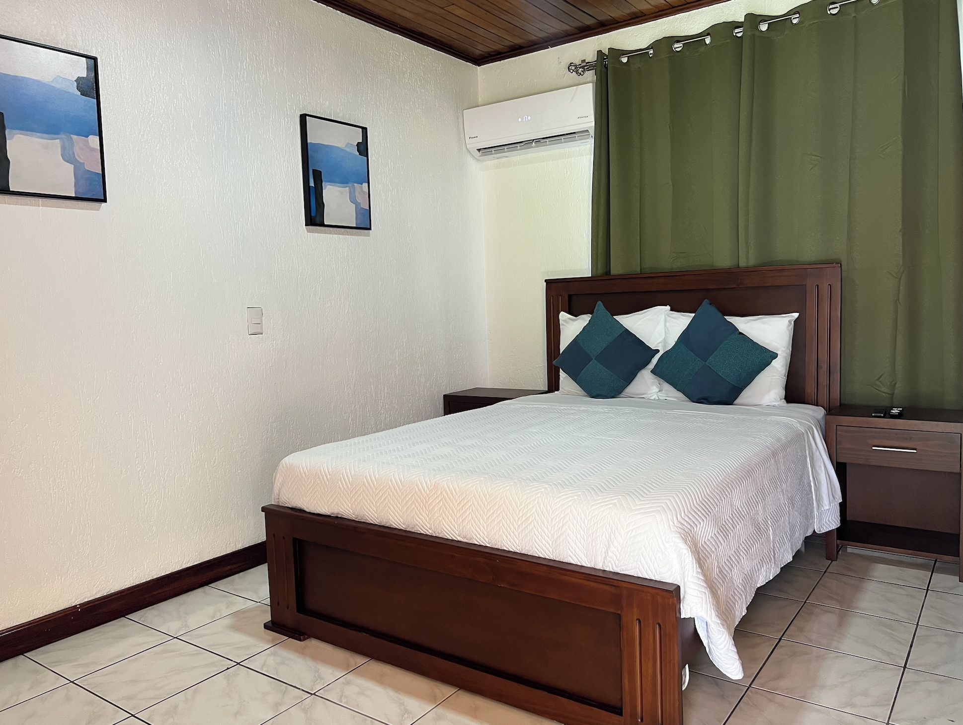 A bedroom with a wooden bed, white bedding, two pillows, a nightstand, a wall-mounted air conditioner, and a green curtain.