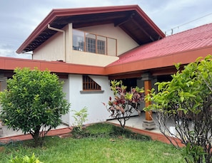 Una casa de dos pisos con techo rojo, balcón y un jardín con diversas plantas.