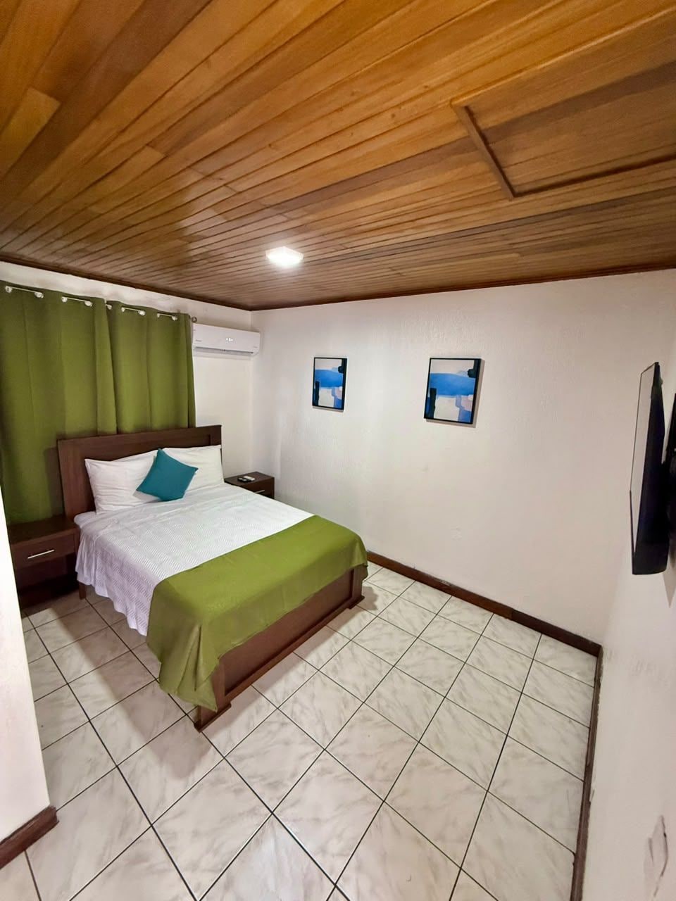 Un dormitorio con techo de madera, una cama con colcha verde, dos mesitas de noche y un aire acondicionado instalado en la pared.