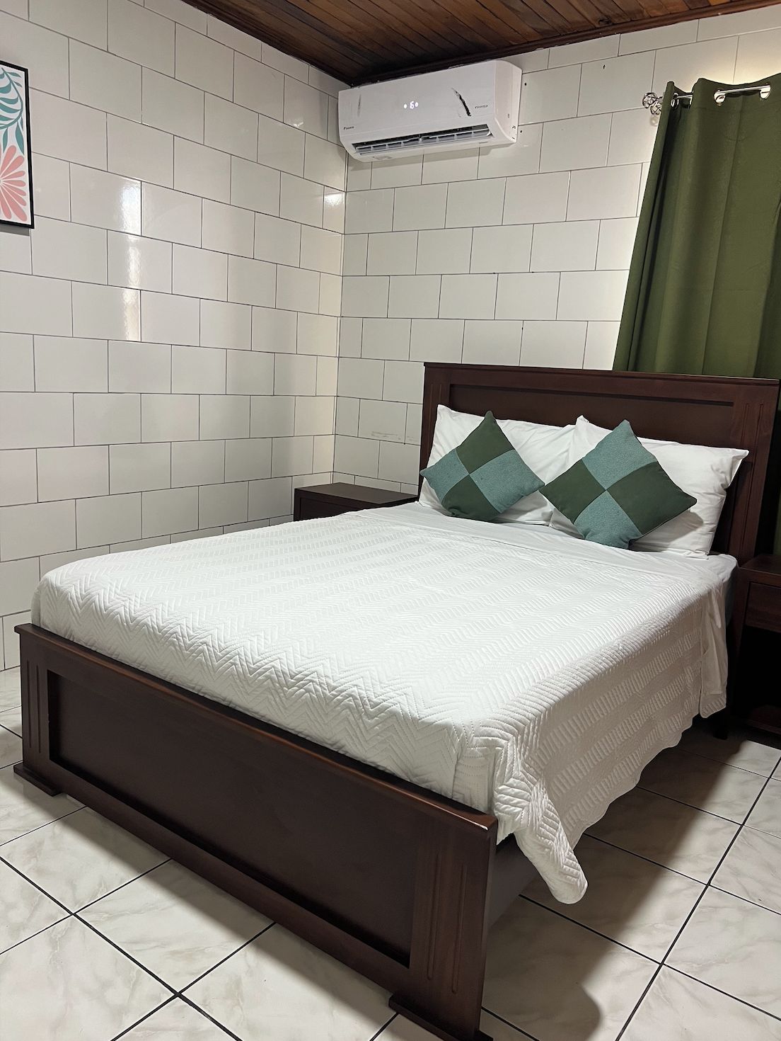 A neatly made bed with a wooden frame, white bedding, and two green pillows. An air conditioning unit is mounted on the wall above the bed.