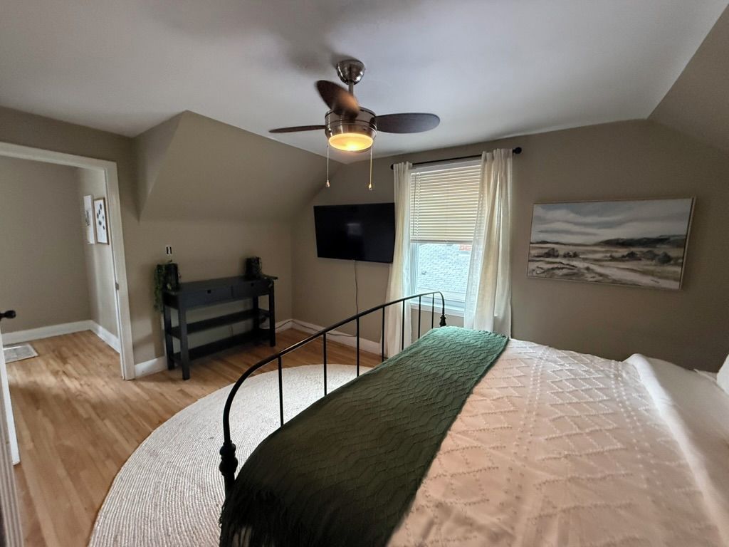 Une chambre à coucher avec un lit, un ventilateur de plafond, une télévision, un bureau et une fenêtre avec des rideaux.