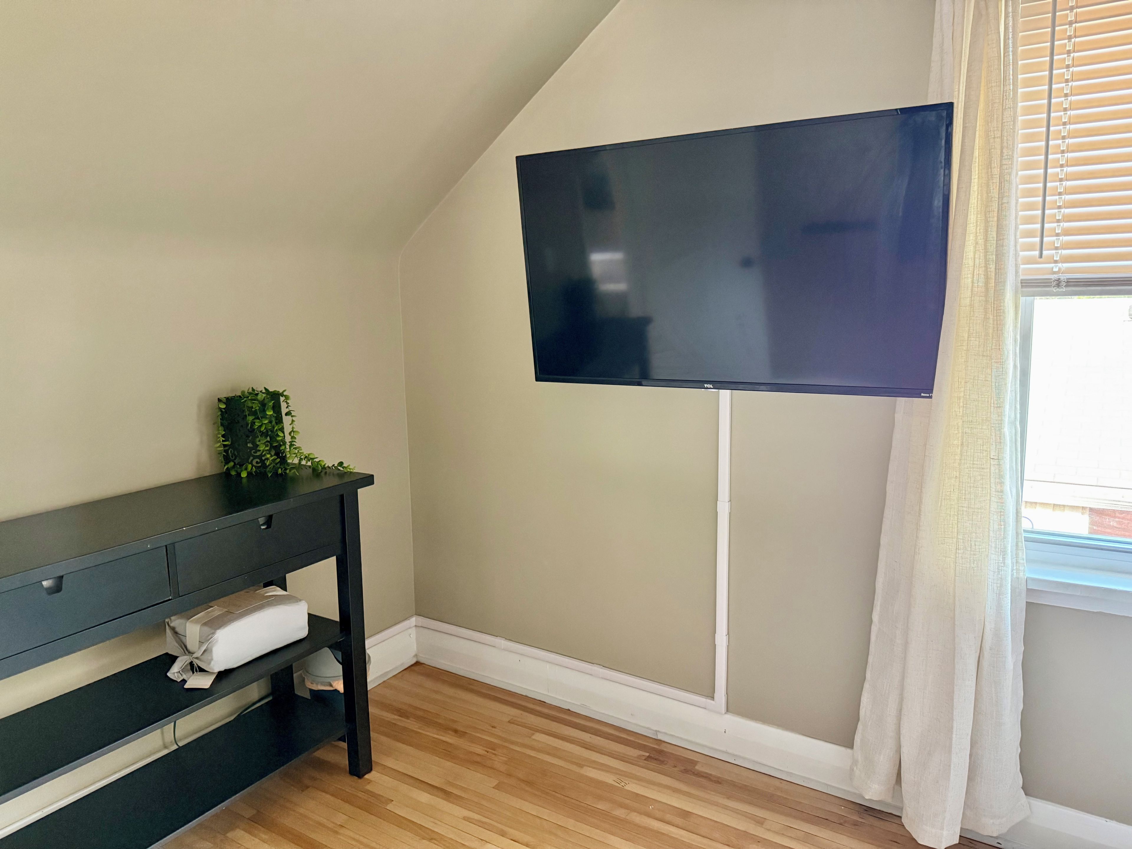 Ein Zimmer mit einem an der Wand befestigten Flachbildfernseher, einem dunklen Holzkonsole mit Pflanze und verpacktem Gegenstand sowie einem Fenster mit Jalousien.