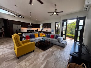 A modern living room with a sectional sofa, a yellow armchair, and a coffee table. There is a kitchen in the background with dark cabinetry and a dining area. - Tropical Escape 3 BR Private Pool / lawn / Fenced (Sosua)
