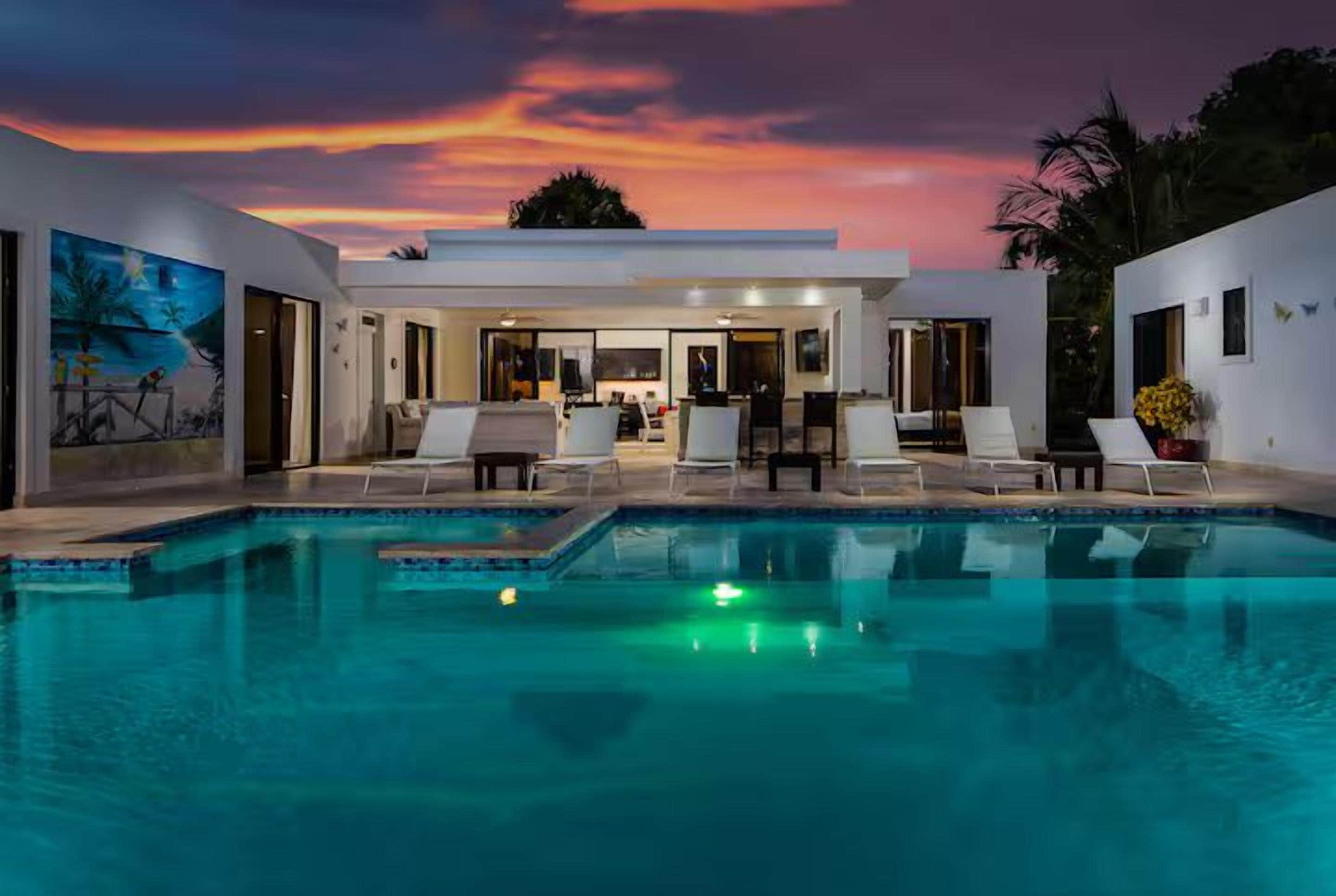 A swimming pool with lounge chairs, a dining area, and a sunset view.