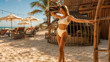 Escena de playa con sillones de descanso y sombrillas, una estructura de bambú y cielo despejado.