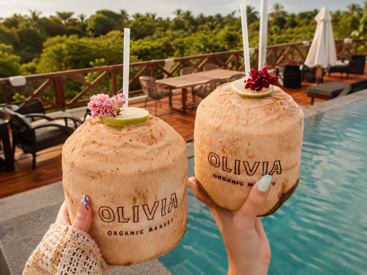 Two coconut drinks with 'OLIVIA ORGANIC MARKET' branding, held by individuals with a pool and outdoor seating in the background.