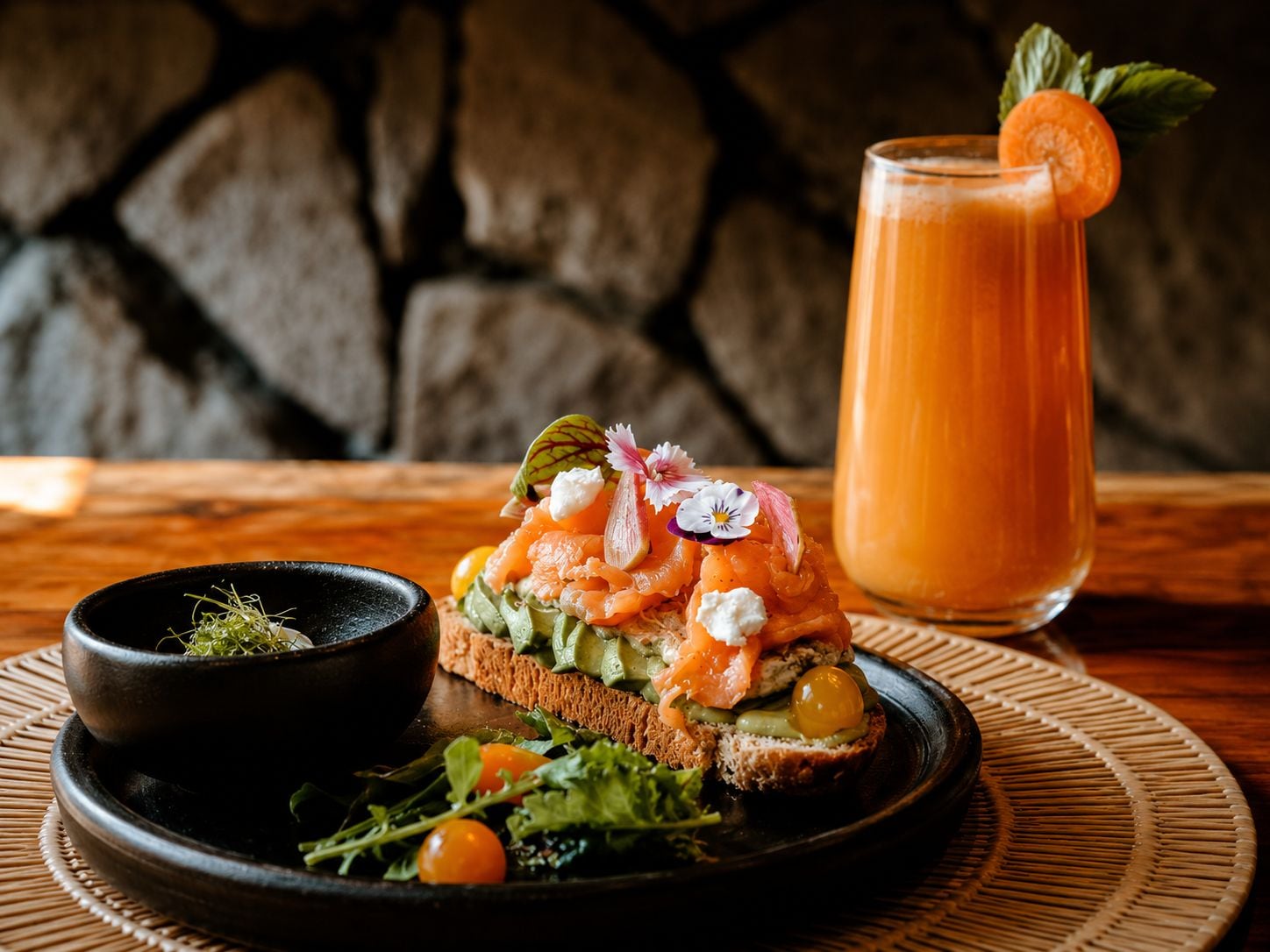 Un plato con salmón ahumado, queso crema y vegetales sobre una rebanada de pan, acompañado de un vaso de jugo de naranja y un tazón pequeño de vegetales de hoja verde.