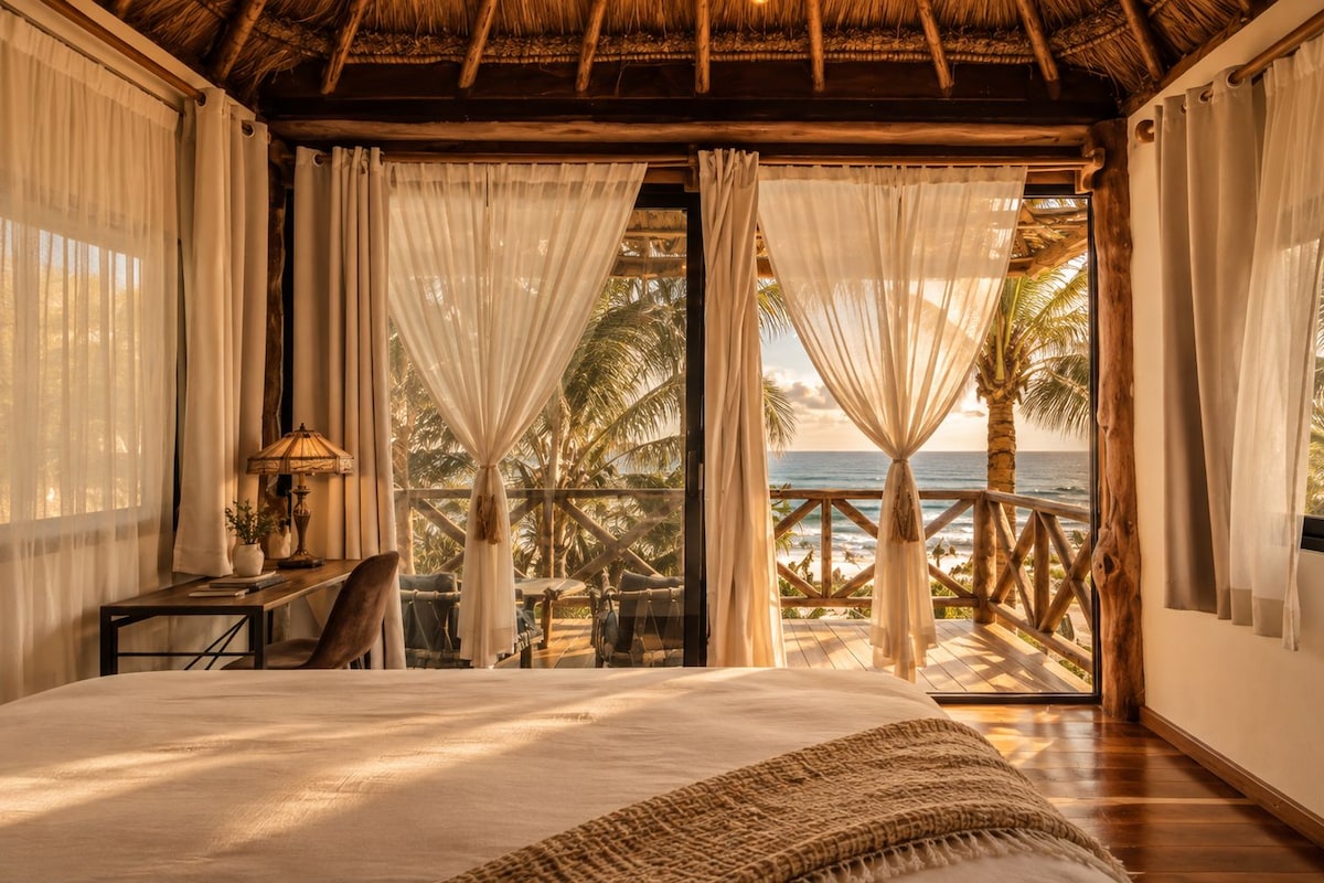 A bedroom with a large bed, a desk with a lamp, and a view of the ocean through curtains.