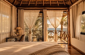 A bedroom with a large bed, a desk with a lamp, and a view of the ocean through curtains.