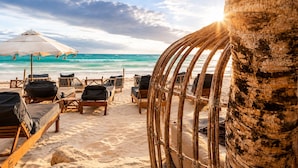 Una playa con sillas reclinables de madera y una sombrilla grande, una palmera y el océano al fondo.
