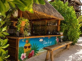 Un bar tiki con techo de paja, rodeado de plantas tropicales y un mural con temática playera.