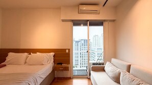 Ett hotellrum med en säng, en soffa, ett fönster med utsikt över byggnader och en luftkonditioneringsanläggning.