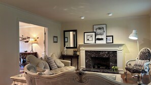 A living room with a fireplace, a sofa, a chair, and a dining area with a table and lamp. - Song • Pinecrest Manor | Forest & Pond (Surrey)