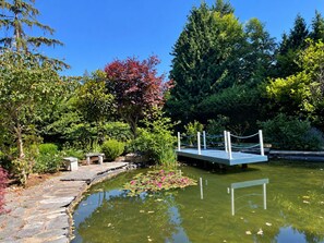A tranquil garden with a stone pathway, a wooden dock, and a pond with lily pads. - Song • Pinecrest Manor | Forest & Pond (Surrey)