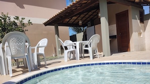 A swimming pool with clear blue water, surrounded by white plastic chairs and a covered patio area. - Casa Brisa De Arraial (Porto Seguro)