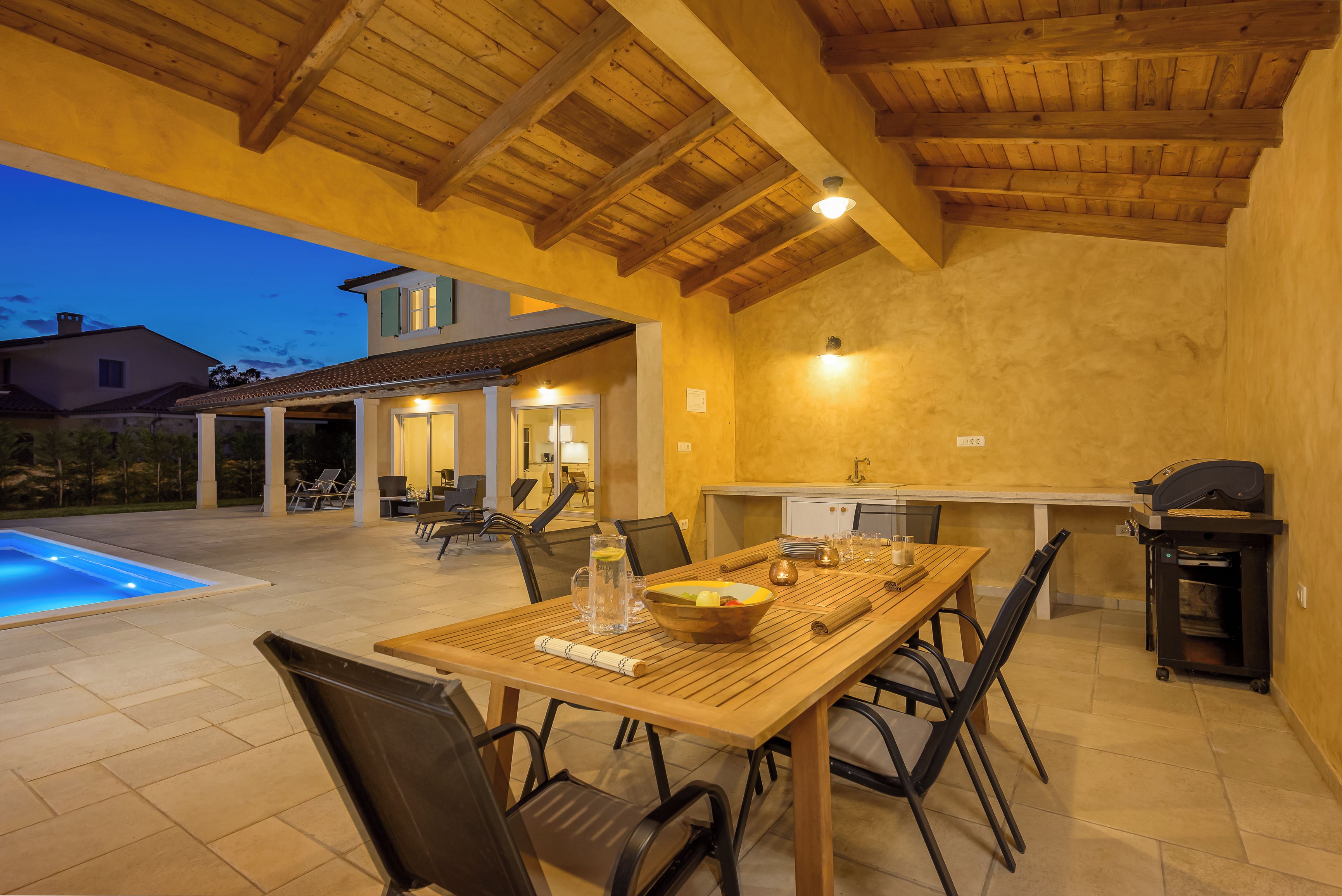 An outdoor dining area with a pool, wooden tables, and chairs, surrounded by a covered patio with a wooden ceiling.