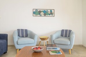 Un salon avec deux fauteuils bleu clair, une table basse en bois, un saladier rempli de fruits et une petite table d’appoint avec des livres.