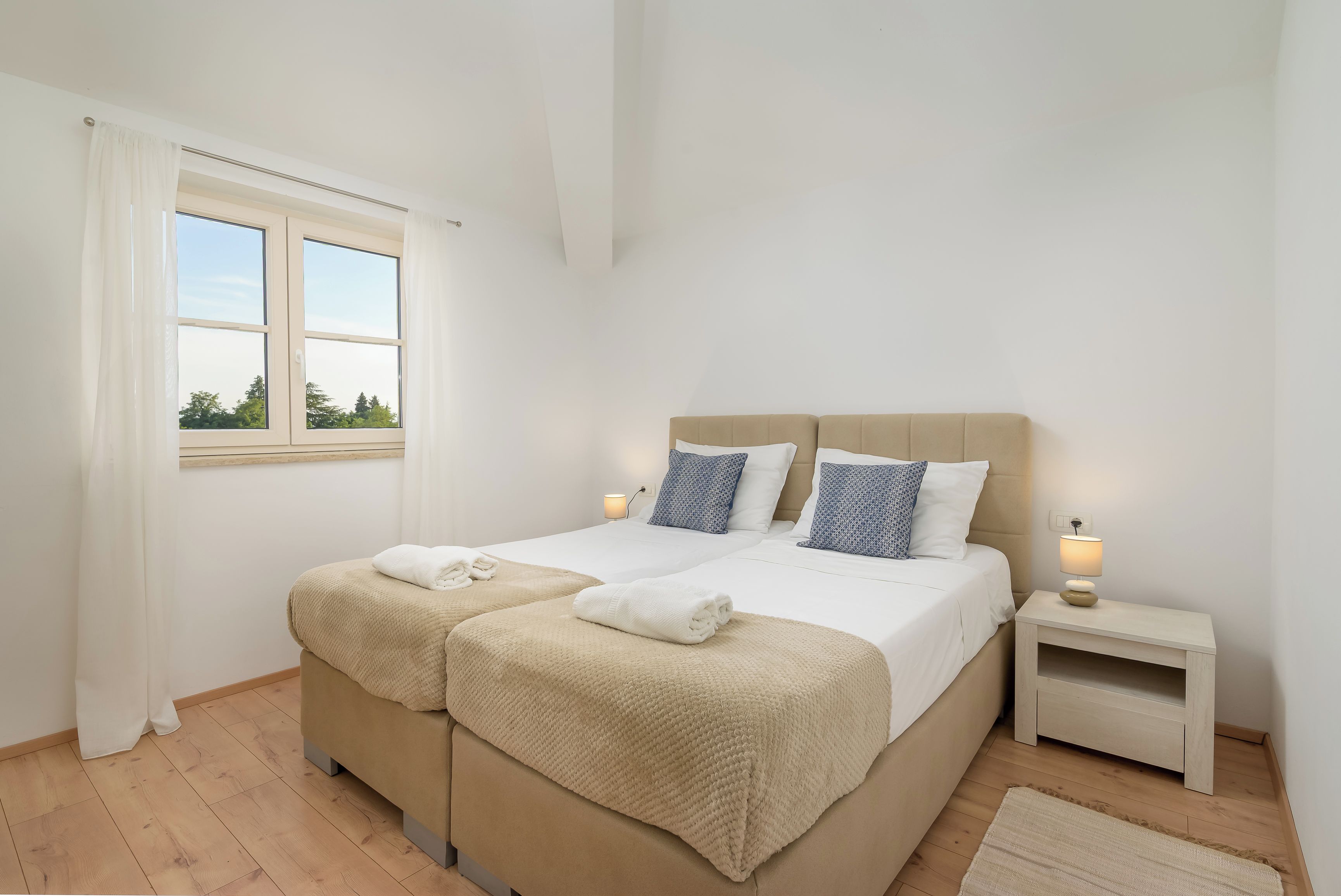 Une chambre à coucher avec un lit double, deux tables de chevet, une fenêtre avec des rideaux et une vue sur les arbres.