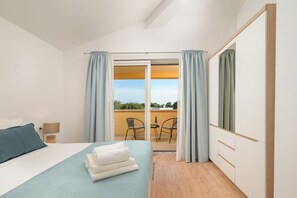Une chambre à coucher comprenant un lit, une table de chevet, une lampe, une fenêtre avec des rideaux et donnant sur un balcon extérieur.