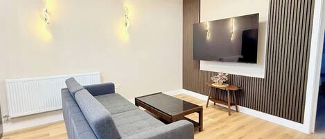 A modern living room with a grey sofa, a coffee table, a flat-screen TV mounted on a wall with vertical paneling, and a radiator.