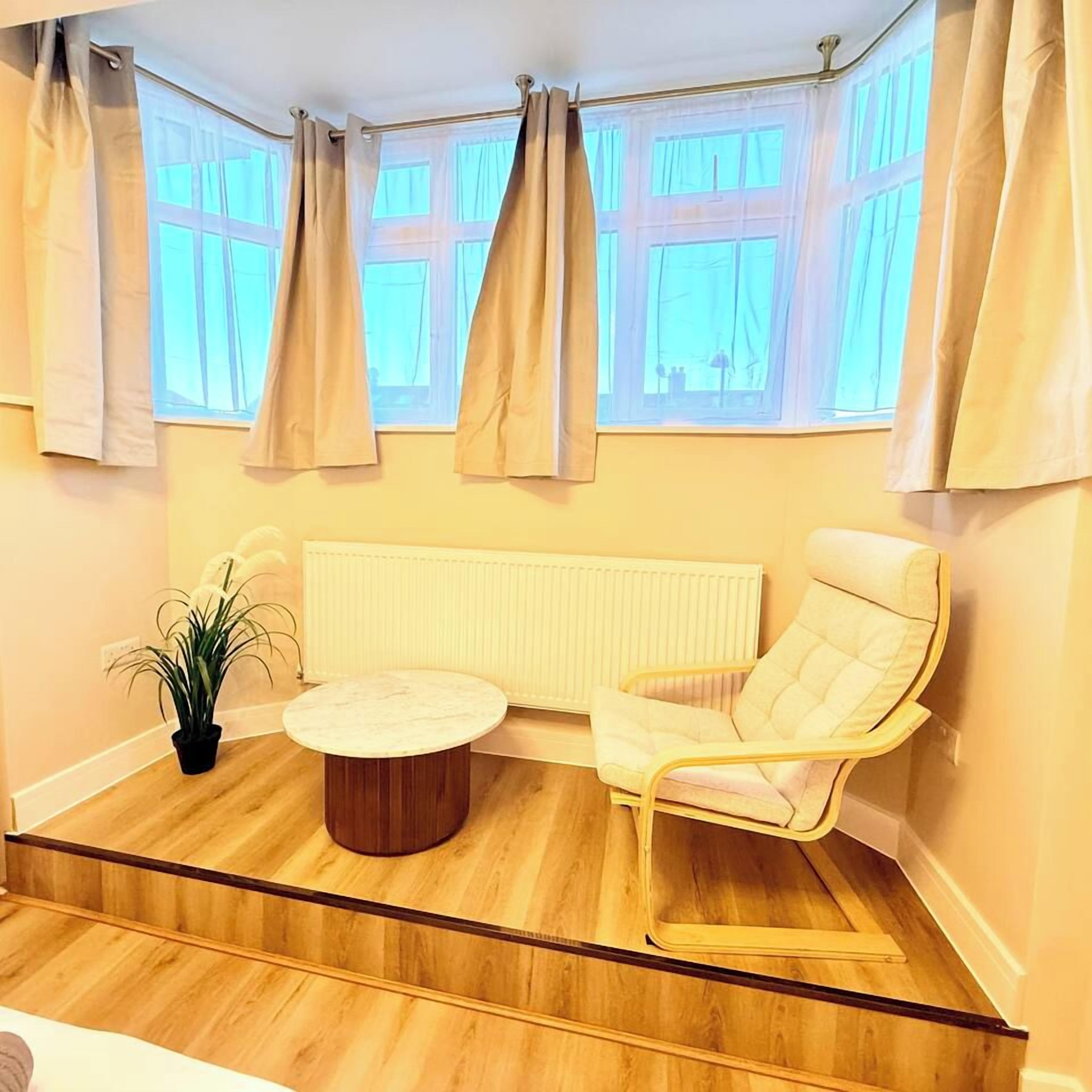 A corner room with a wooden floor, a round table, a cushioned chair, a potted plant, and a radiator.