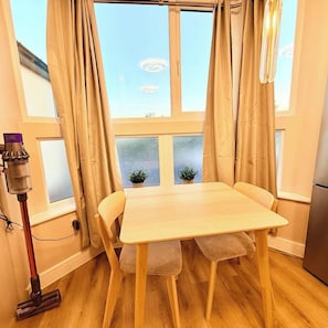 A room with a wooden table and chairs, a window with curtains, a vacuum cleaner, and a small plant on the windowsill. - Chic London Escape | 5 Min Walk to Tube (Greater London)