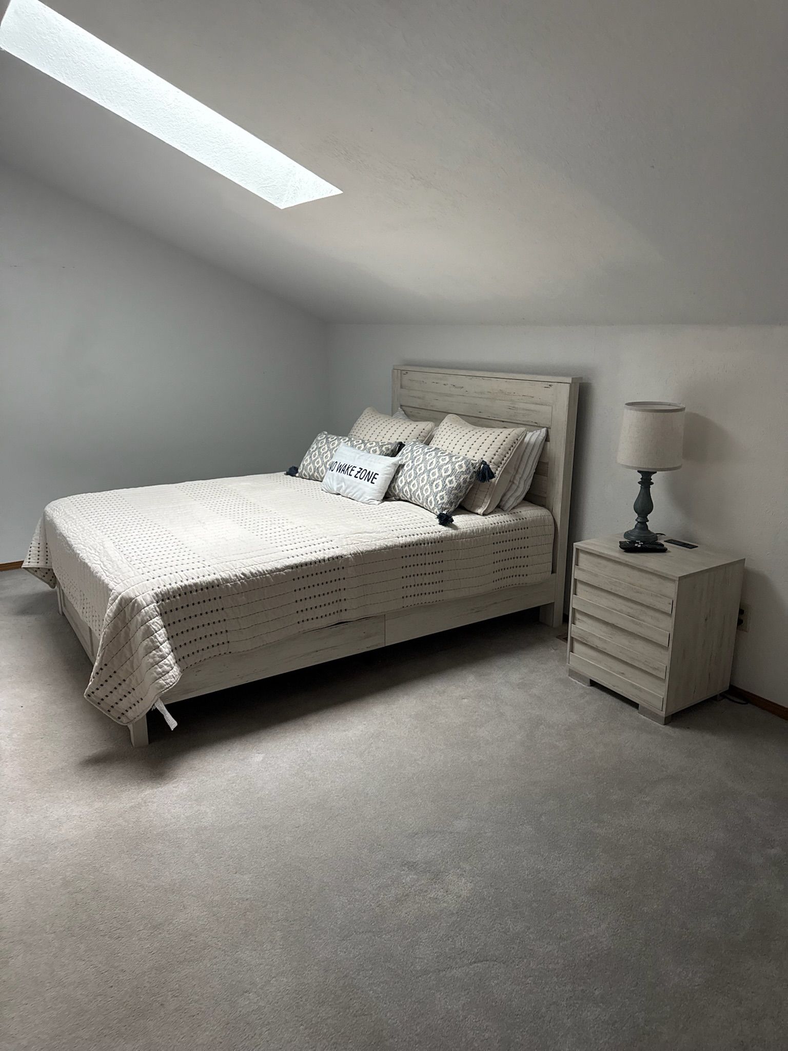 A bedroom with a bed, pillows, a nightstand, a lamp, and a skylight.