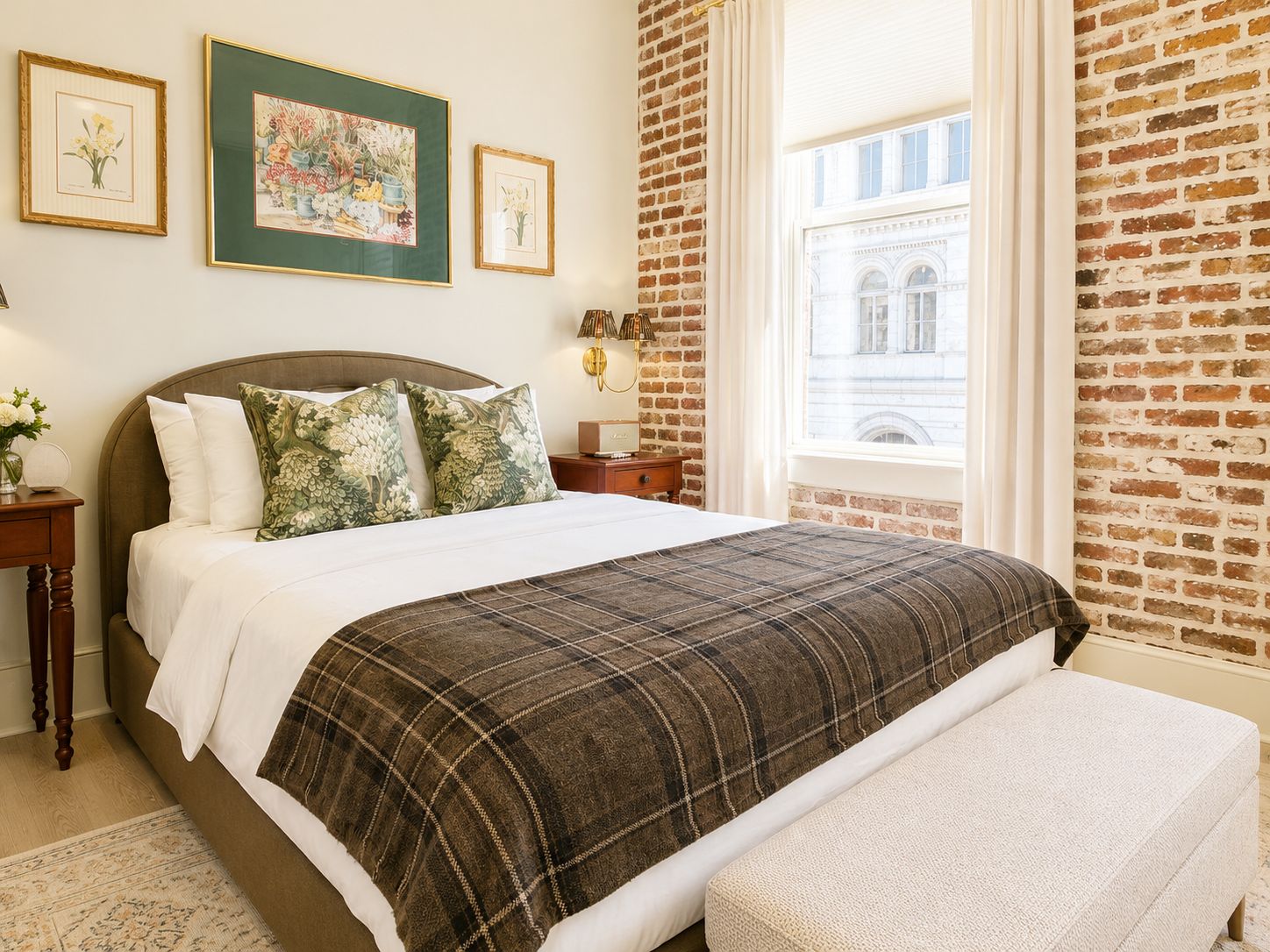 A hotel room with a brick wall, a large bed with a plaid bedspread, a bench, and framed botanical prints on the wall.