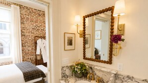 Una camera d'albergo con un letto, bagno con lavandino in marmo e dettagli dorati, e una parete in mattoni.