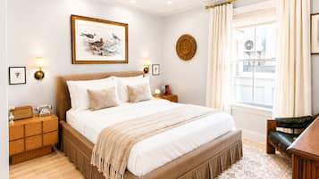A bedroom with a large bed, bedside tables, a nightstand, a lamp, a framed picture, a window with curtains, and a chair.