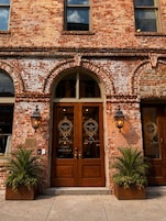 A brick building with a wooden double door entrance, two potted plants, and two wall-mounted lanterns.