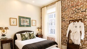 Camera d'albergo con un letto grande, una panca, un comodino, uno specchio con accappatoi e una parete in mattoni.