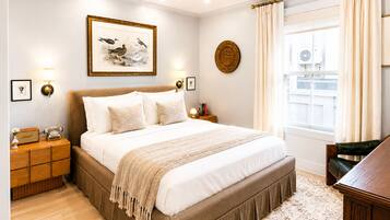 Un dormitorio con una cama grande, una mesita de noche con teléfono, una lámpara de pared, un cuadro enmarcado y una ventana con cortinas.
